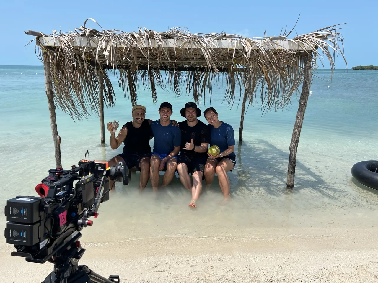Mike and three crew members seated beneath a thatched shelter in the water, with a camera in the foreground